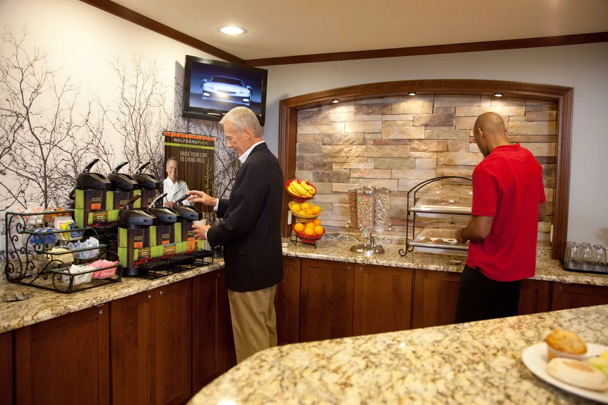 A bright and communal kitchen space showcasing a coffee station, a vibrant fruit display, various breakfast items, and a TV screen. This area is arranged for easy access and casual gathering, encouraging interaction and convenience.