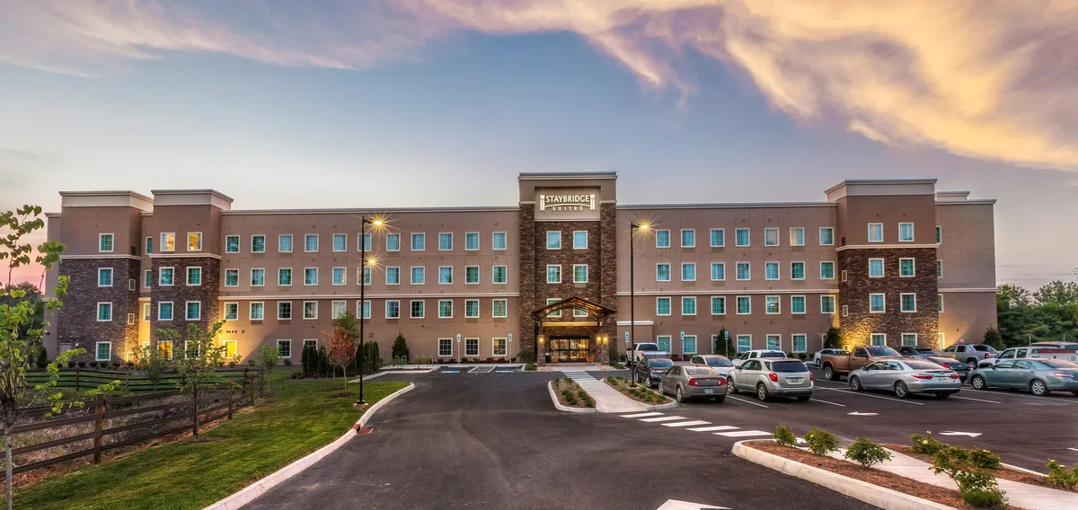 This exterior shot captures the hotel's welcoming facade at sunset, with guests arriving at the entrance canopy and staff offering warm greetings. The parking area is visible, and the scene hints at convenient access for travelers setting out to explore nearby parks and local attractions.