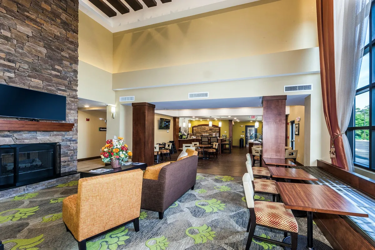 A welcoming interior space featuring a stone fireplace, a cozy seating area, large windows with abundant natural light, decorative flowers, and a dining area set up for gatherings. The arrangement suggests an inviting atmosphere ideal for enjoying meals or socializing.