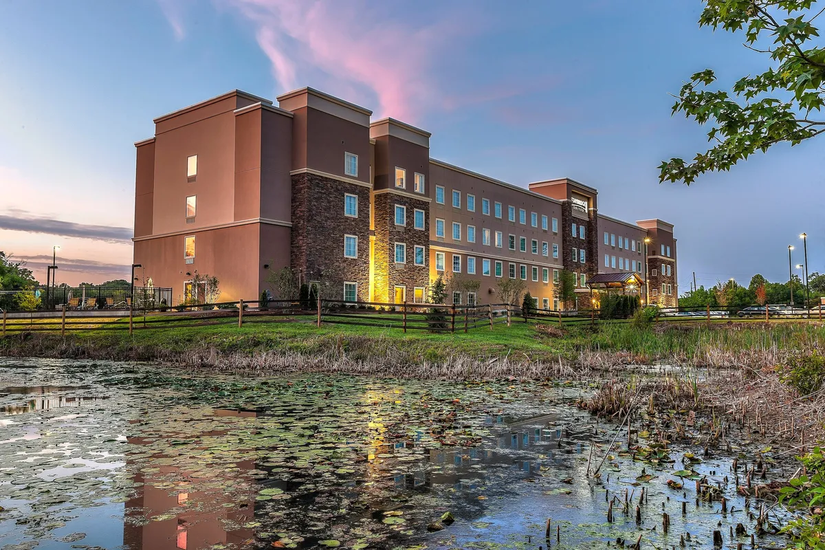 This image captures the hotel building reflected in a tranquil body of water, framed by lush greenery and set beneath a vivid sunset sky. The outdoor environment invites guests to enjoy relaxing moments by the water, evening strolls, or simply take in the scenic views.